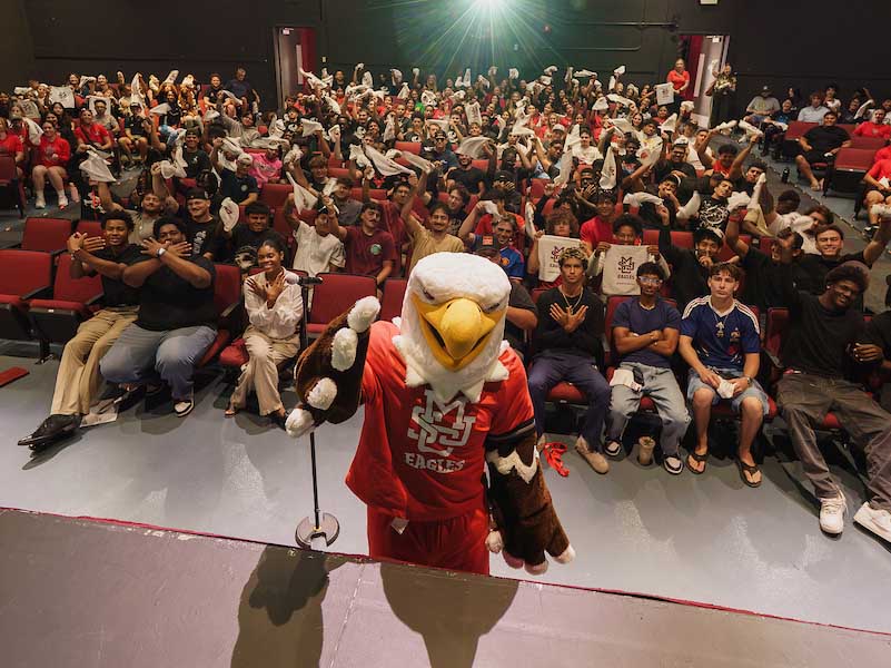An eagle mascot stands on a stage in front of a large audience seated in an auditorium, many holding up white items.