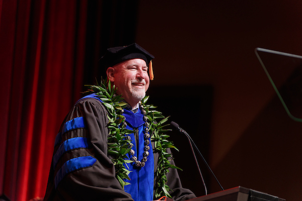A person in academic regalia, adorned with a green lei and a medal, smiles while speaking at a podium during a formal ceremony.